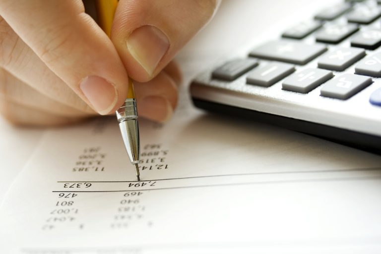 A hand holding a pen, writing on a sheet next to a calculator.